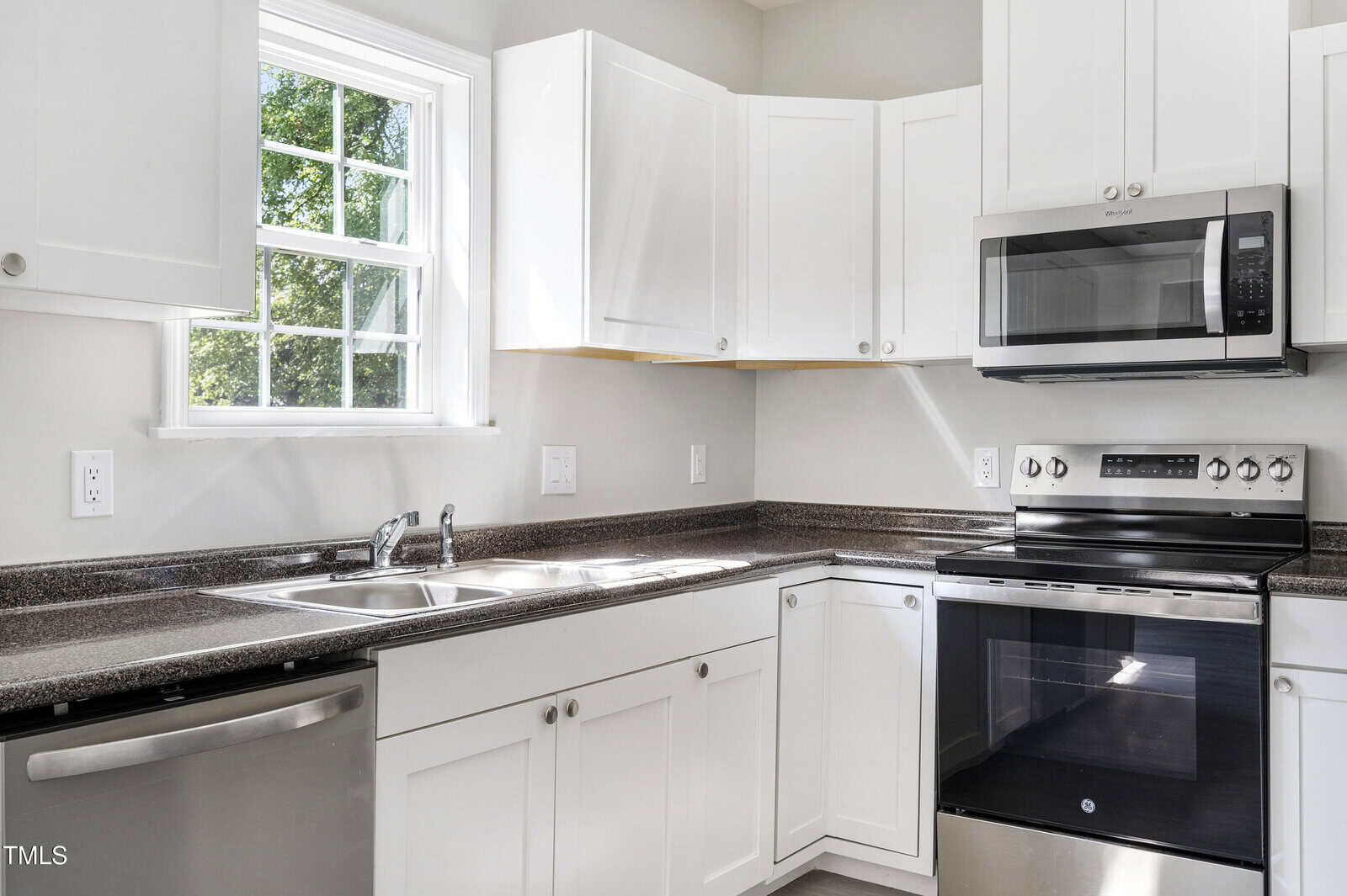 A modern kitchen featuring white cabinets, a stainless steel dishwasher, a double-basin sink, an electric range, and a microwave. A window to the left allows natural light to illuminate the space. The countertops are dark and contrast with the white cabinets.