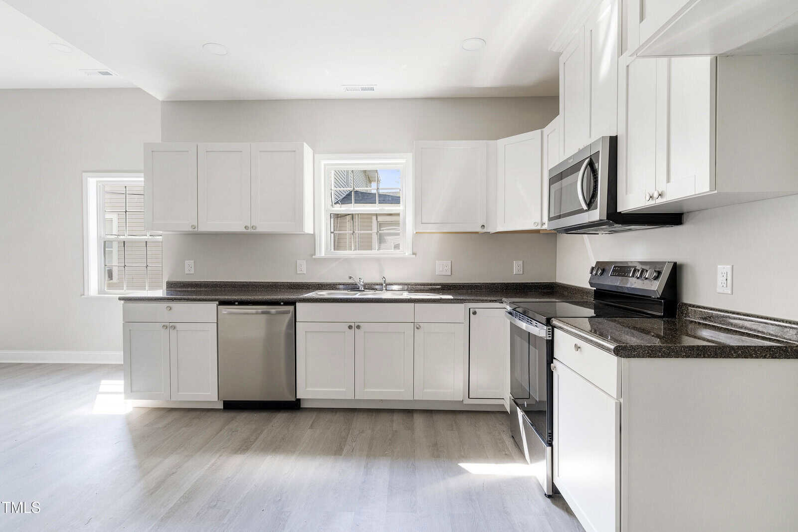A modern kitchen with white cabinetry and dark countertops. It features a stainless steel dishwasher, oven, and microwave. There is a double sink under a window with natural light illuminating the space. The floor is light-colored wood.