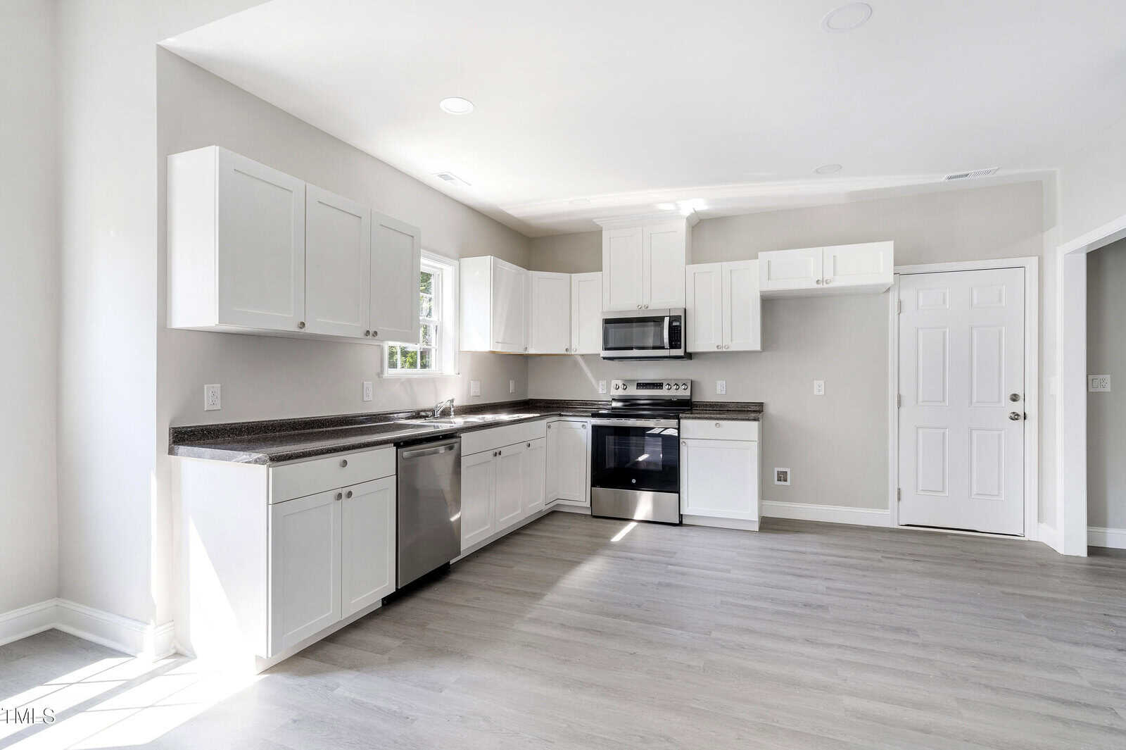 A modern kitchen with light gray walls and flooring, featuring white cabinets, dark countertops, a stainless steel dishwasher, oven, and microwave. A window lets in natural light, and a white door is visible on the right.