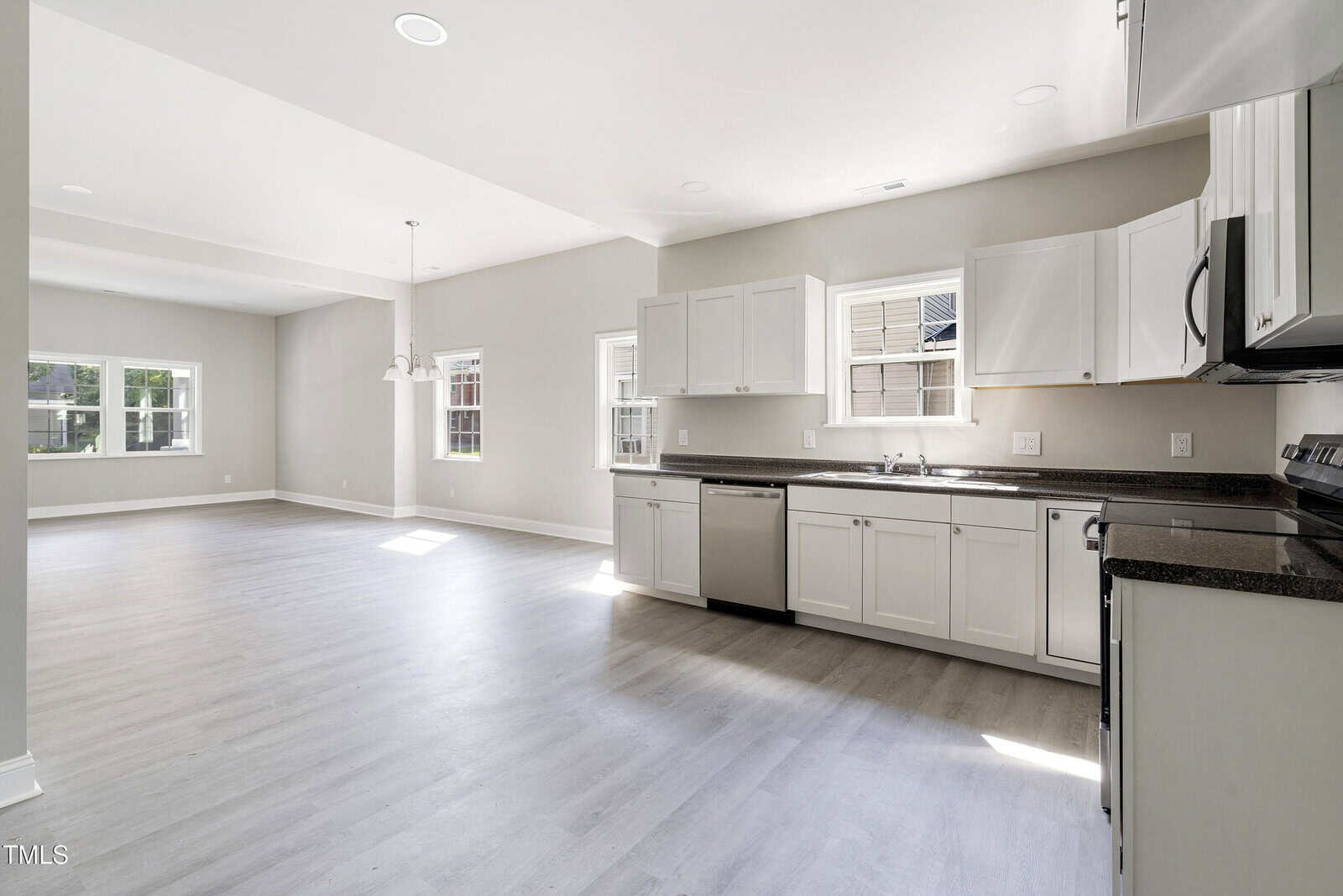 A spacious, empty kitchen and dining area with light-colored walls and flooring. The kitchen features white cabinets, a dark countertop, a stainless steel dishwasher, and stove. Large windows provide natural light to the open space.