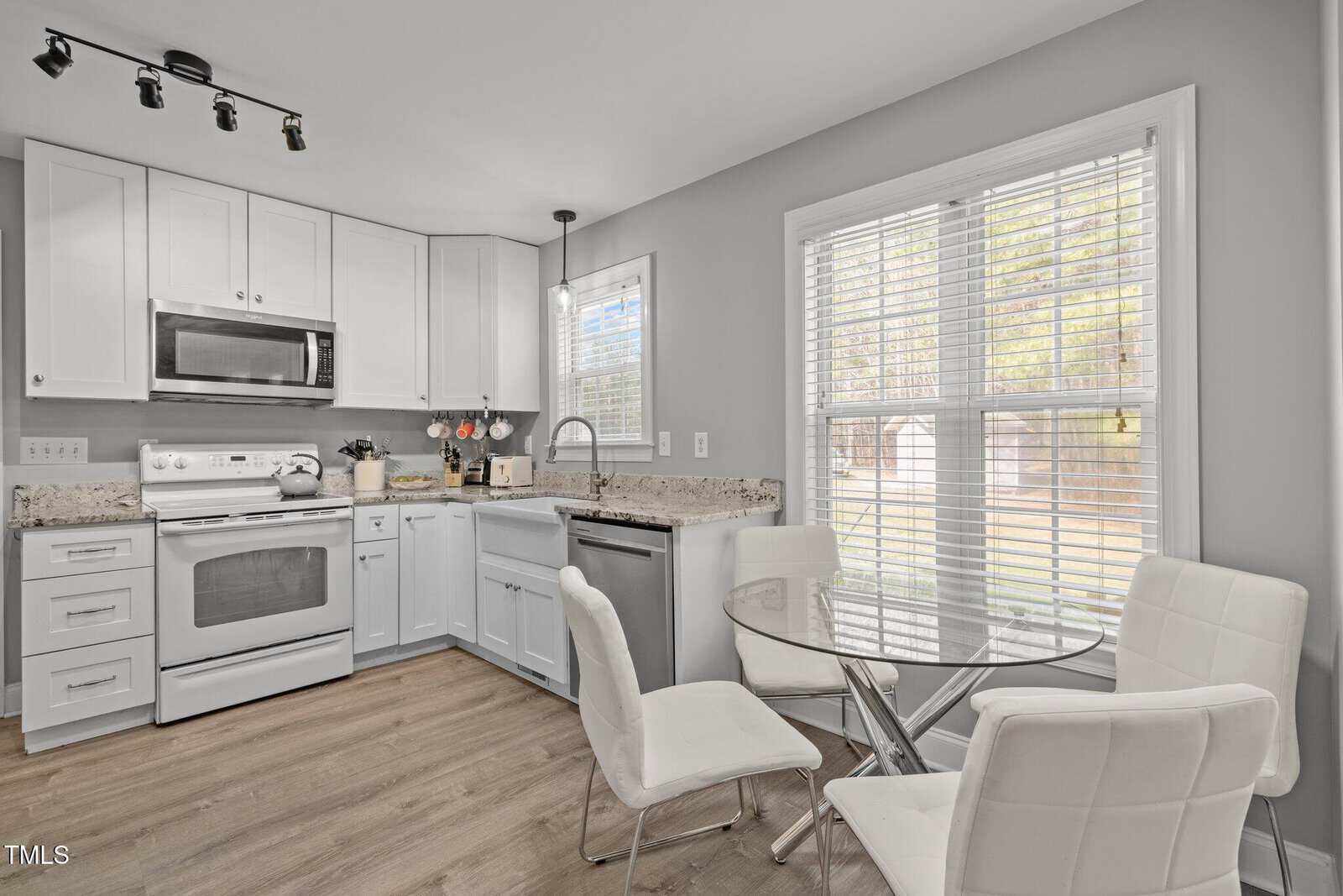 Modern kitchen with white cabinets, granite countertops, stainless steel appliances, and a small round glass dining table with four white chairs next to a large window with blinds. Light wood flooring throughout.