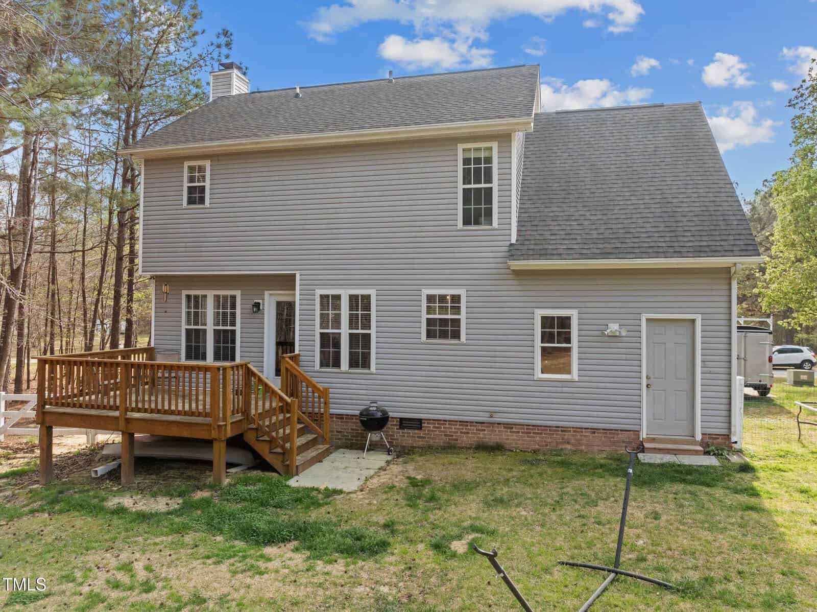 A two-story house with gray siding, multiple windows, and a shingled roof. There is a small wooden deck with stairs, a grill on a stone patio, a back door, and a grassy yard with trees in the background.