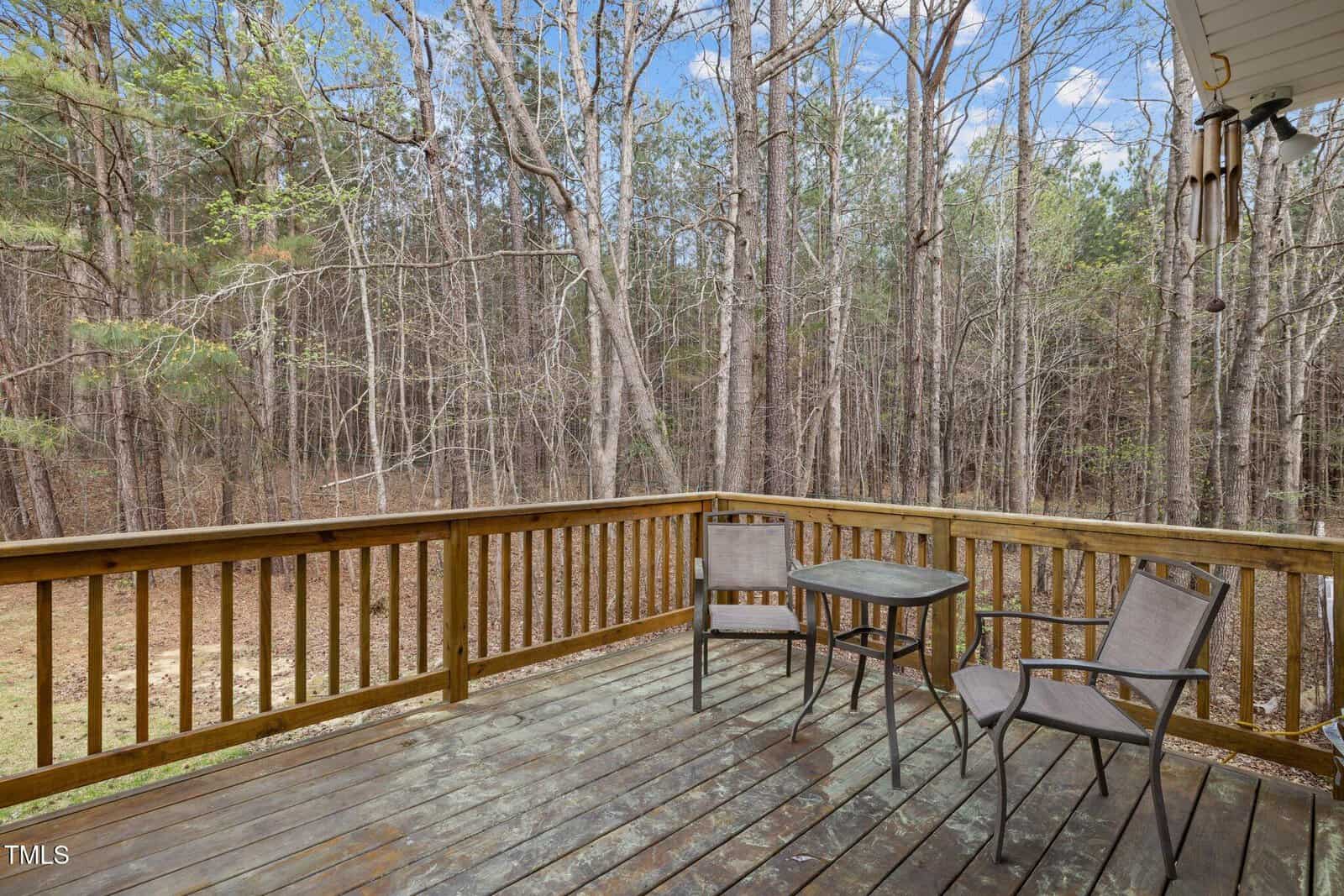 A wooden deck with a small table and three chairs overlooks a wooded area with tall trees and a partly cloudy sky in the background. A wind chime hangs on the right side near the roof.