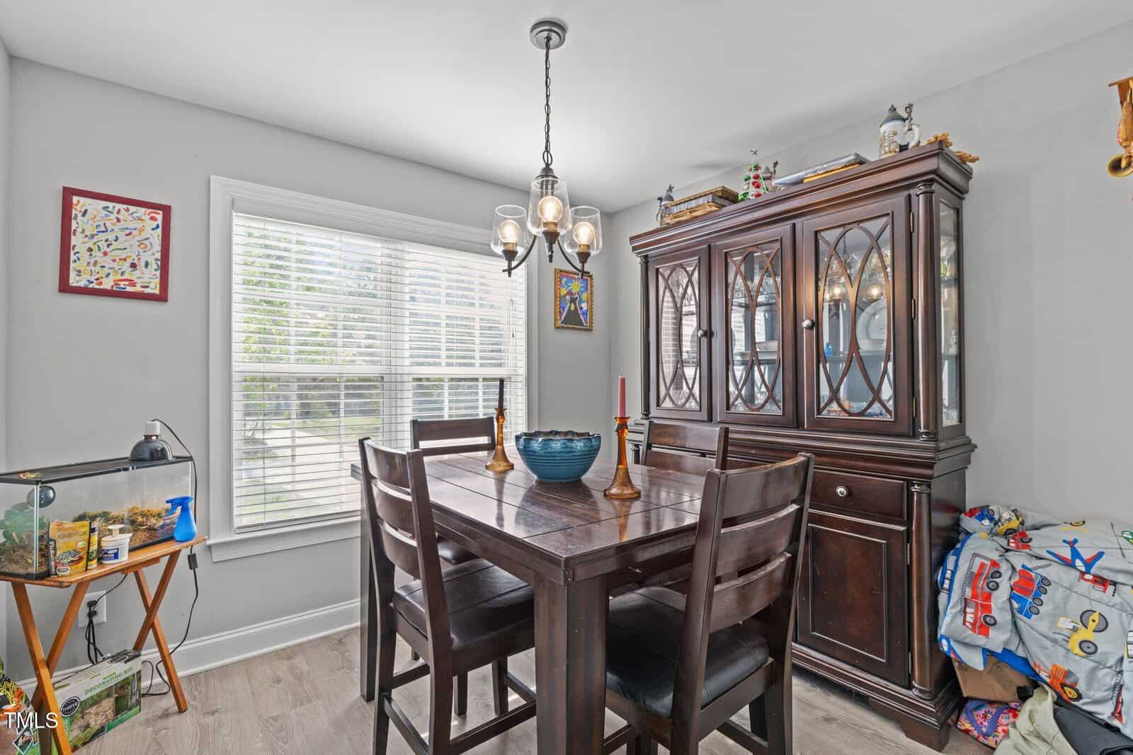 A dining room with a dark wood table and four chairs, a china cabinet, a window with blinds, framed art, a fish tank on a side table, and children’s toys in the corner. The floor is light wood.
