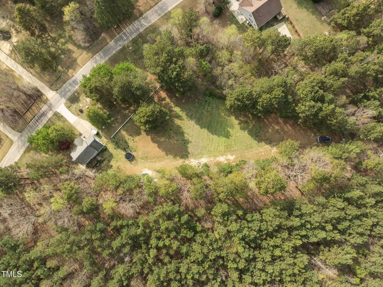 Aerial view of a residential area with a house, driveway, and large, grassy yard surrounded by dense trees. A road borders the property, and other houses are partially visible nearby.