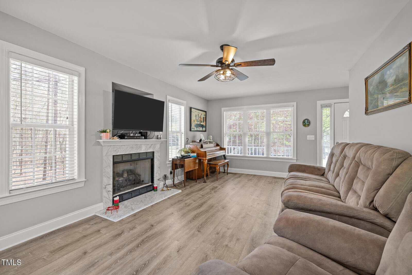 A living room with wood flooring, a brown reclining sofa, a fireplace with a TV above it, a wooden piano, large windows with white blinds, and a ceiling fan with a light fixture. Walls are painted light gray.