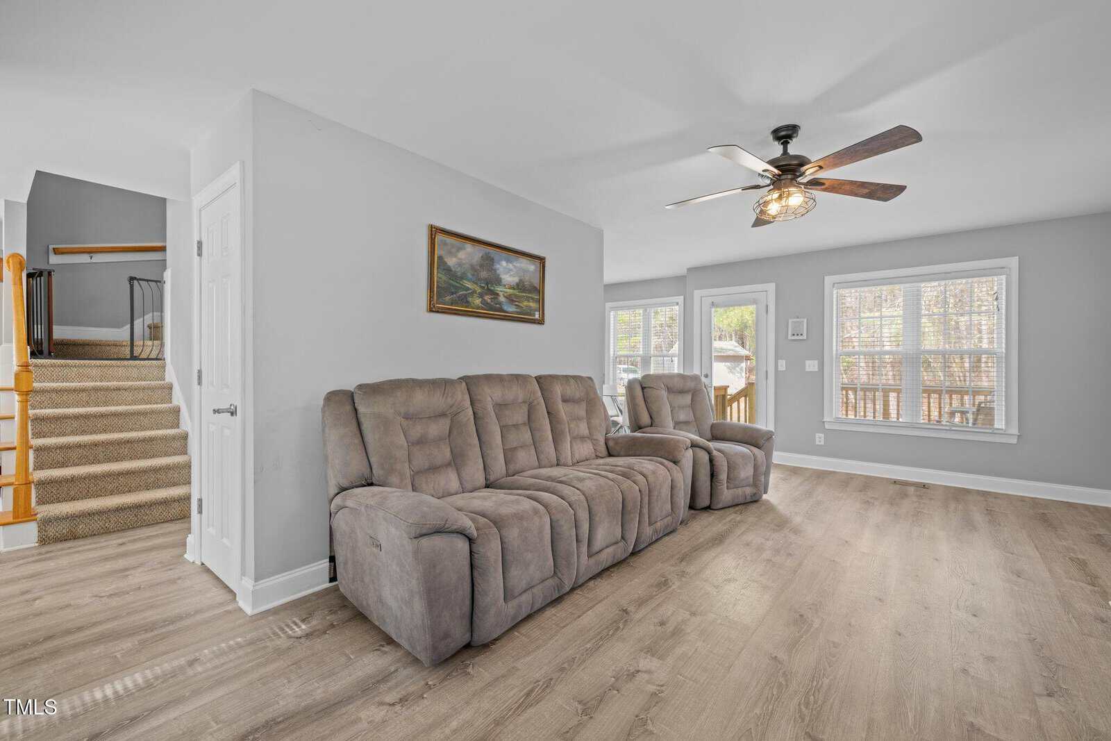 Living room with gray reclining sofa, wood-look flooring, ceiling fan with light, two large windows, stairs with beige carpet and wooden railing, and a landscape painting on a light gray wall.
