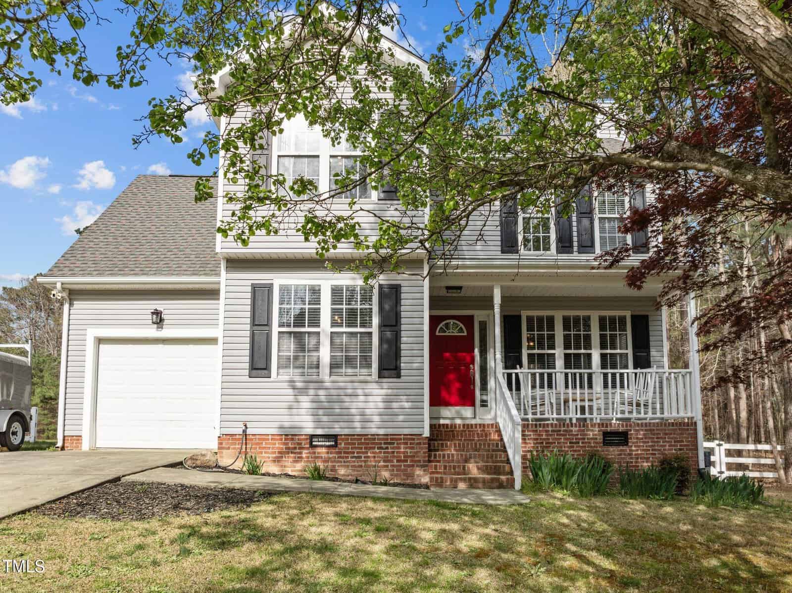 Two-story house with gray siding, white trim, and black shutters. The front door is red, and there is a covered porch with white railings. The house has an attached garage and a driveway, with trees in the yard.