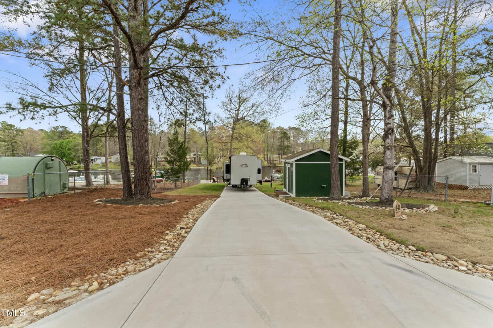 A long concrete driveway leads to a white RV parked between a green shed on the right and a fenced area on the left, surrounded by tall trees and mulch beds; lake and houses are visible in the background.