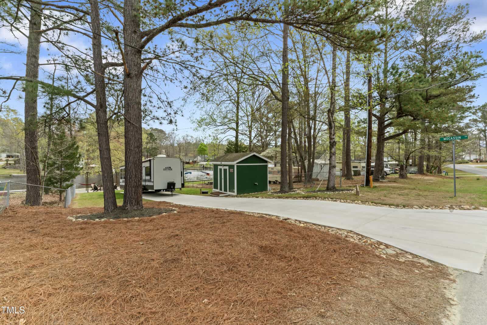 A paved driveway leads to a green shed and a white trailer among tall pine trees in a wooded area, with a road and residential homes visible in the background under a clear sky.