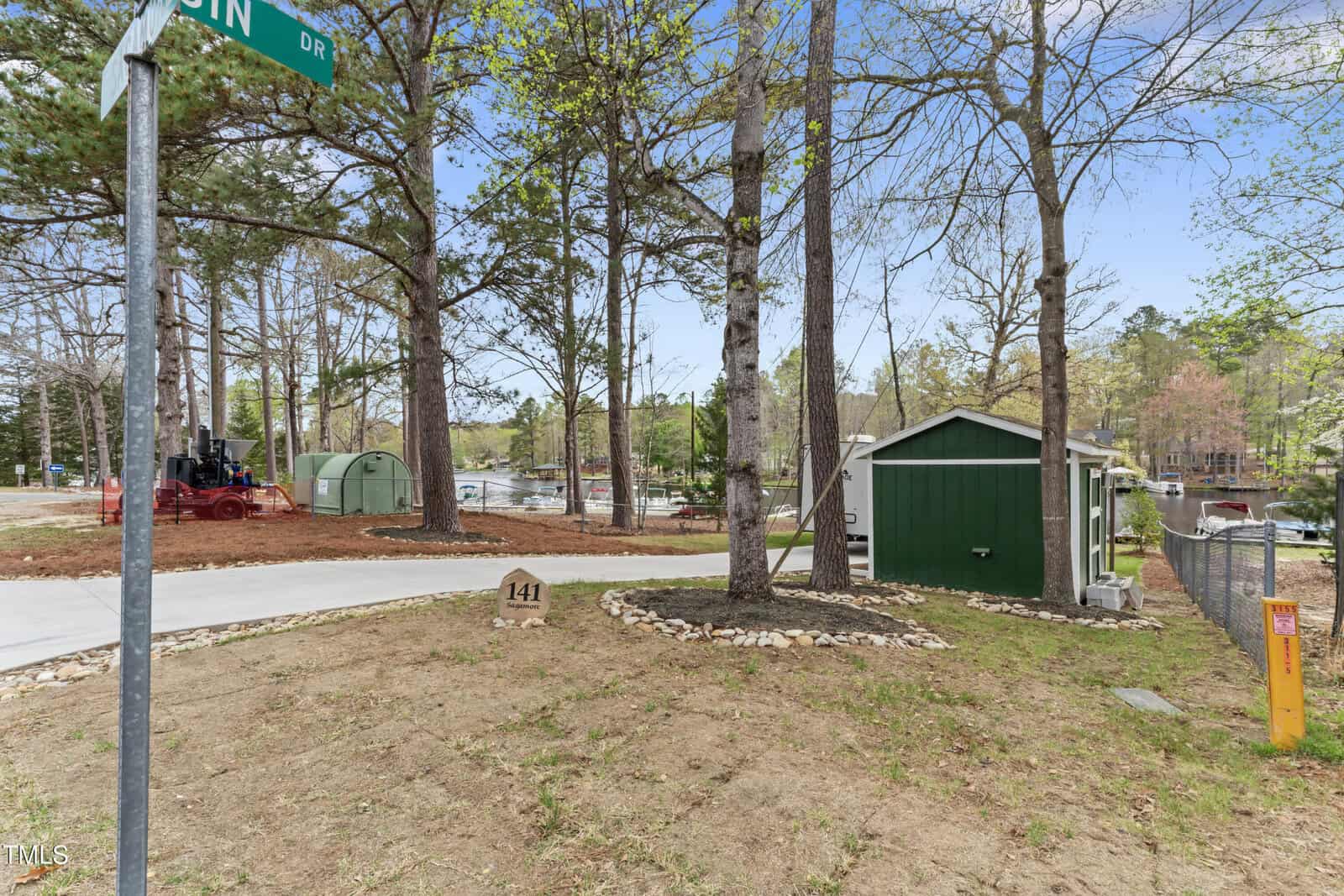 A small green shed sits beside a fenced area and driveway. The yard has sparse grass and several tall trees. A lake with docks, boats, and neighboring houses is visible in the background. A street sign reads “PIN.”.