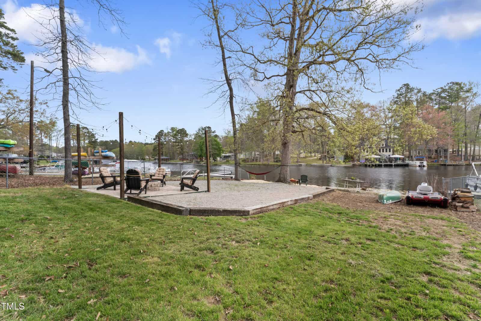 A backyard with a gravel patio area featuring Adirondack chairs around a fire pit, string lights, a hammock, trees, and a view of a calm lake with docks and boats under a partly cloudy sky.