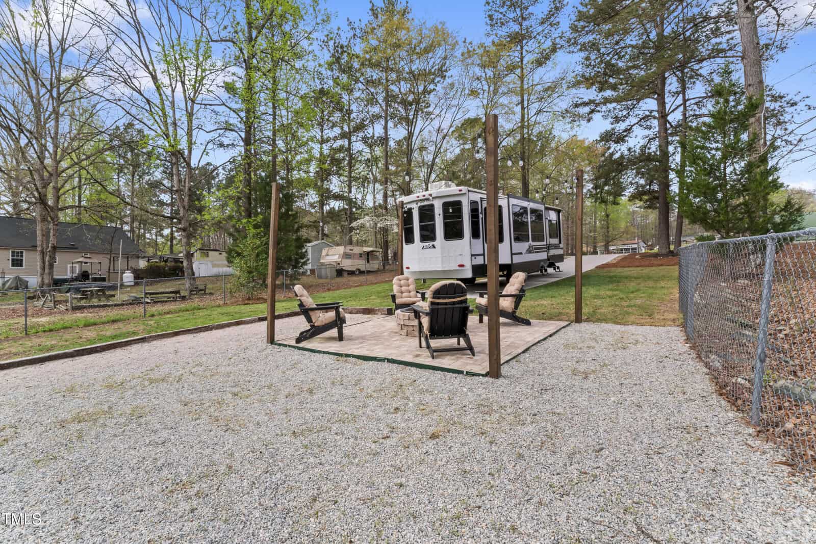 A gravel lot with a paved patio area featuring four chairs around a fire pit. An RV is parked nearby, and trees and houses can be seen in the background, with a chain link fence on the right side.