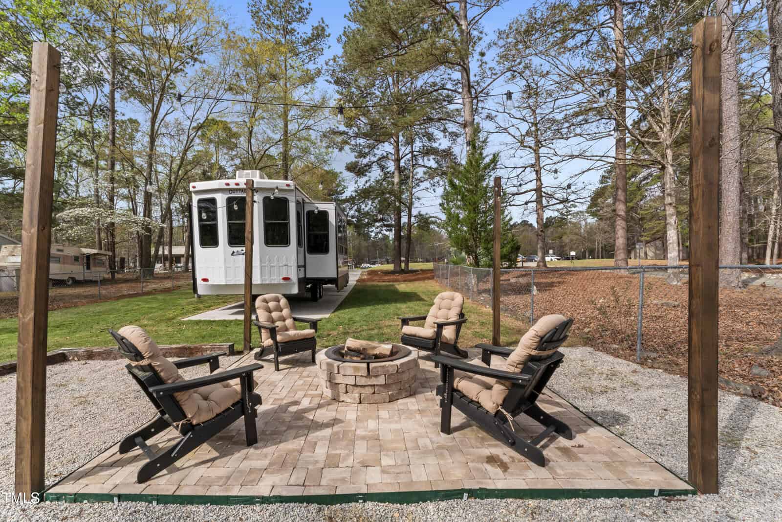 Four cushioned patio chairs are arranged around a stone fire pit on a paved area, with a white RV parked on grass in the background and trees surrounding the scene.