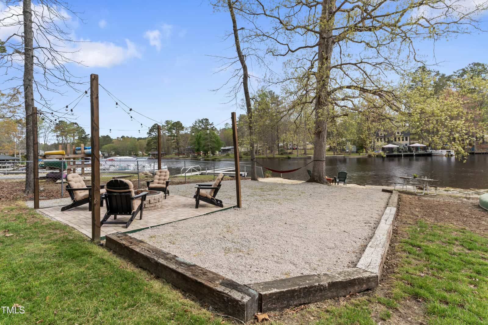 Patio area with four cushioned chairs around a fire pit on gravel, string lights overhead, hammock to the right, and a view of a calm lake with docks and trees in the background.