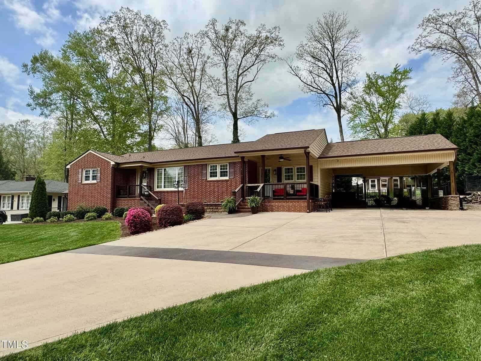 Located at 1802 Greenbriar Road in Henderson, this charming brick single-story house features a covered carport and a large driveway. The home is encircled by a manicured lawn, shrubs, and trees. A cozy porch with chairs graces the front under a partly cloudy sky.