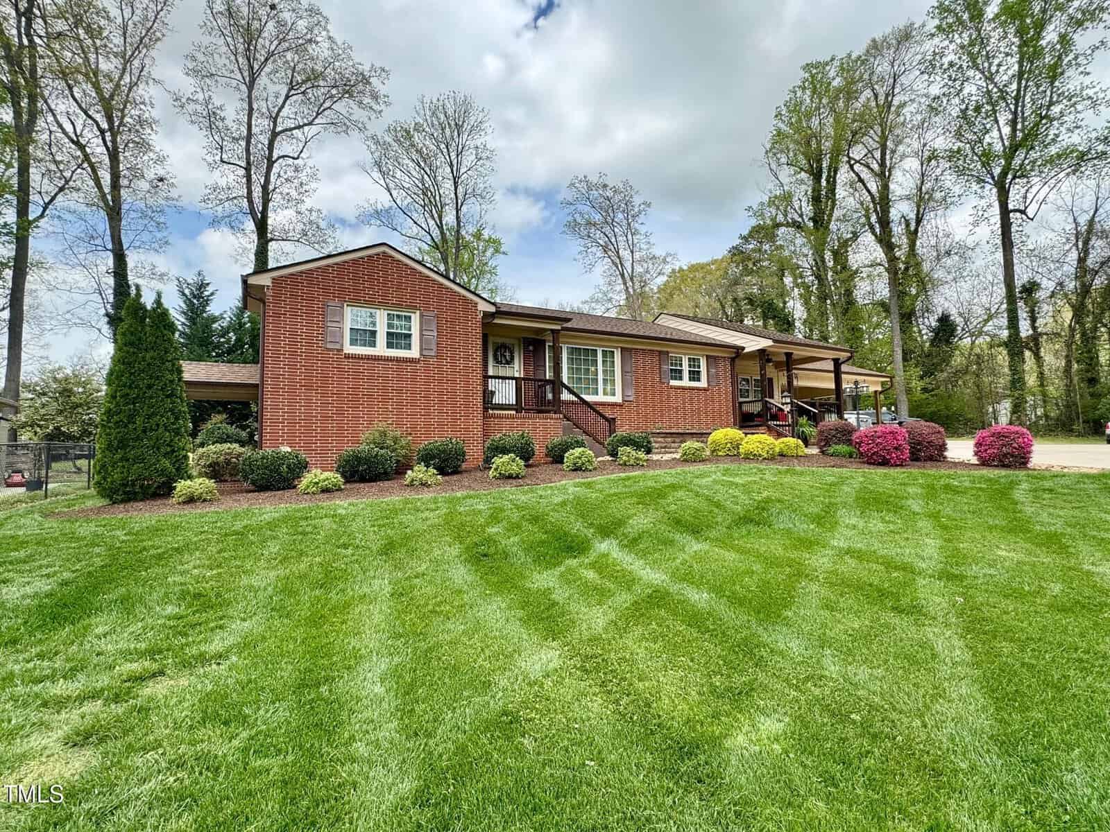 A brick house with brown trim and a covered porch is surrounded by a well-kept lawn. Bushes and small trees line the front, and colorful shrubs add contrast. The sky is partly cloudy, and tall trees border the property in the background.