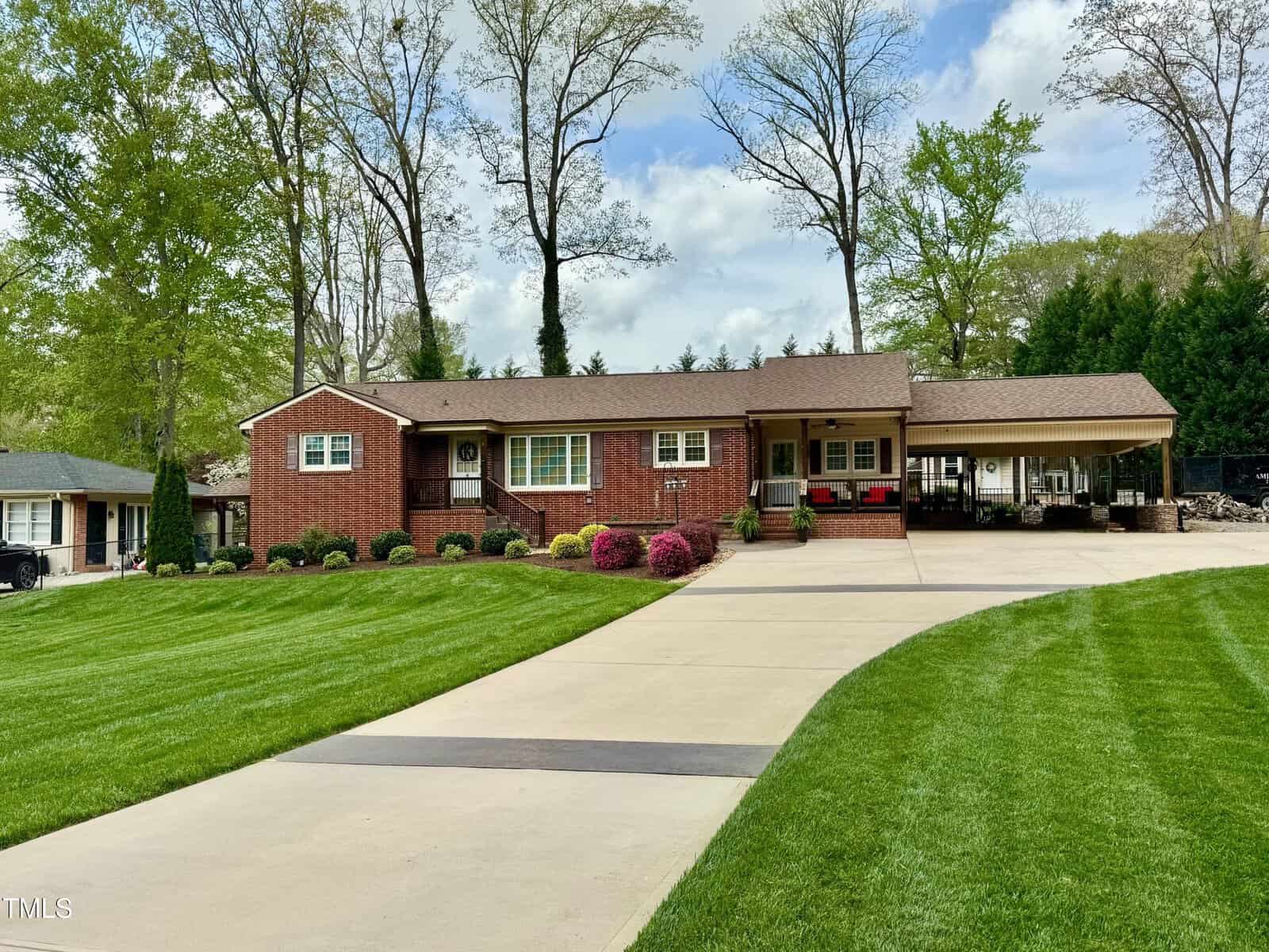 A one-story brick house with a well-maintained lawn and a long concrete driveway. The front features a covered porch with columns. There are bushes with pink flowers lining the front. Tall trees with sparse foliage are in the background.
