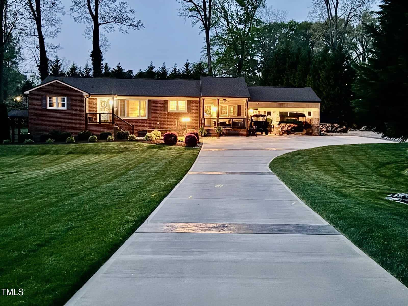 A single-story brick house with a well-maintained lawn and a curved concrete driveway leading up to it. The house is lit from inside, and there are trees in the background, with the sky suggesting it's evening or early morning.