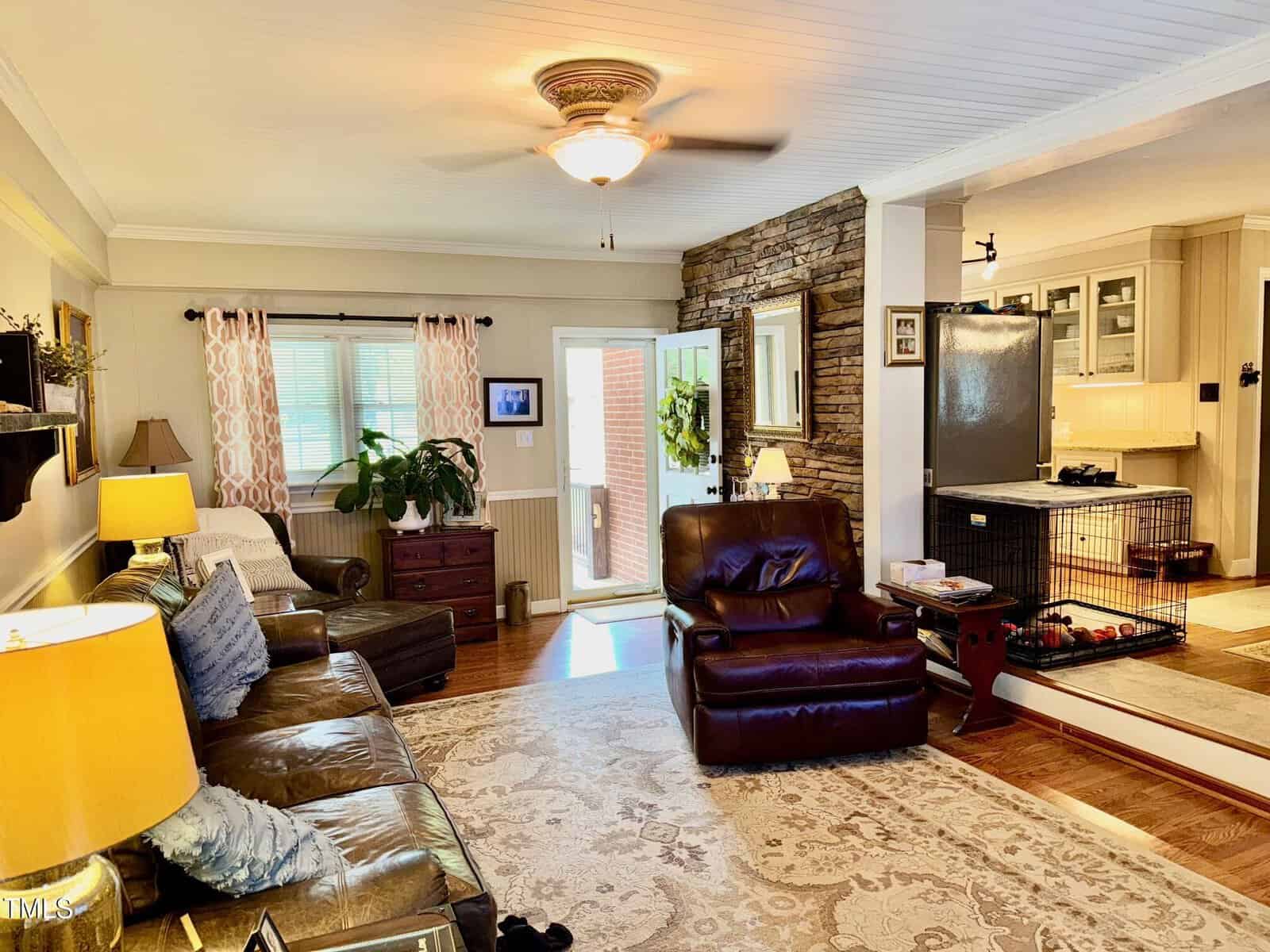 A cozy living room features a brown leather sofa and armchair, a ceiling fan, and a patterned rug. There's a stone accent wall, a window with curtains, and a dog crate near the entrance to a kitchen area with light-colored cabinets.