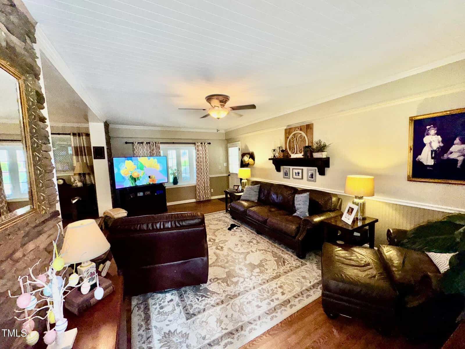 A cozy living room with white paneled ceiling, wooden floor, and a large area rug. It features brown leather sofas, a TV, and decorative items on shelves. A ceiling fan is centered, and a large window allows natural light to enter.