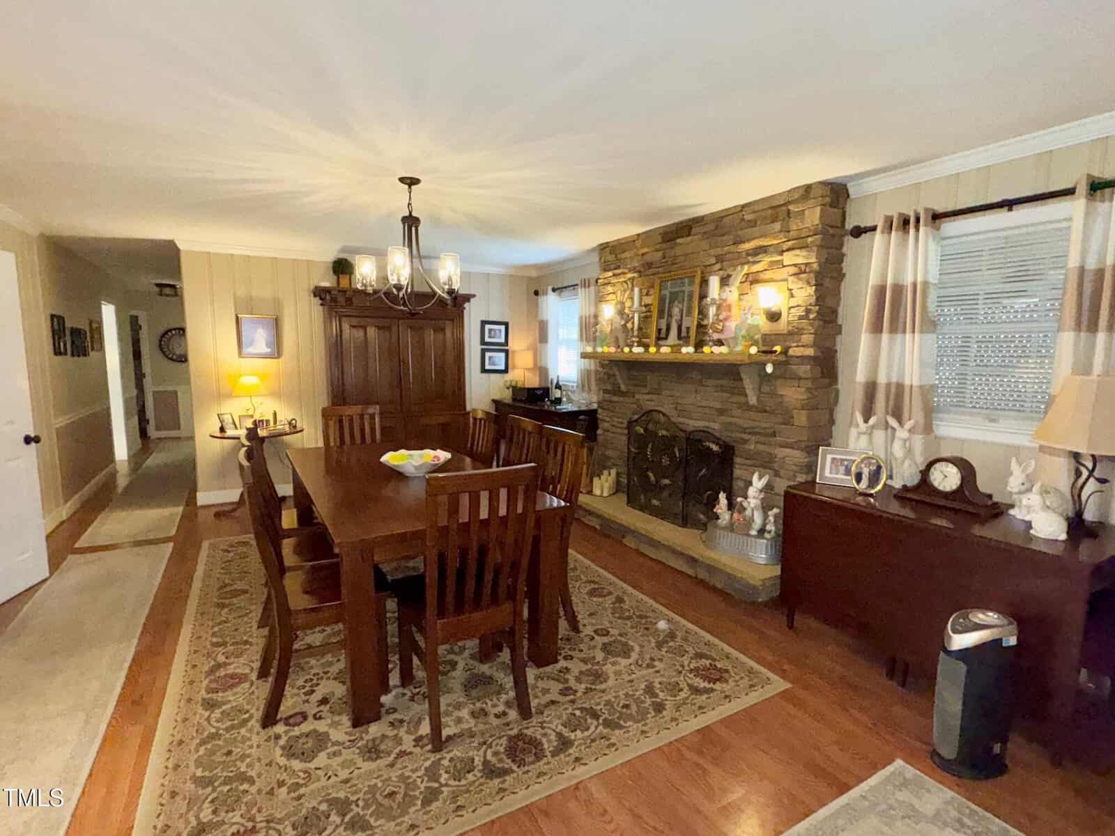 A dining room with a wooden table and six chairs on a patterned rug. A stone fireplace with a wrought iron screen is on the right, adorned with various decorations. A chandelier hangs above the table, and framed pictures are on the walls.