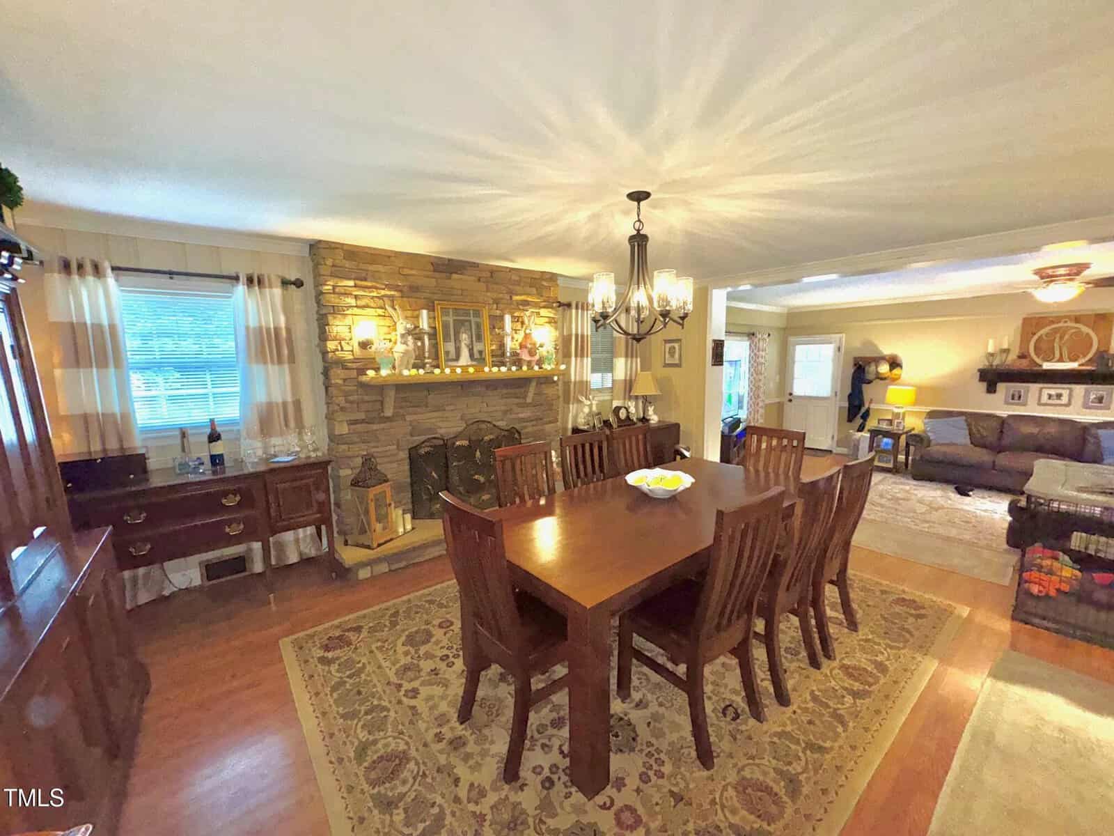 A dining room features a wooden table with eight chairs on a patterned rug, centered under a chandelier. A brick fireplace with a mantel is on an adjacent wall. The room opens into a living area with a couch and additional decor elements.