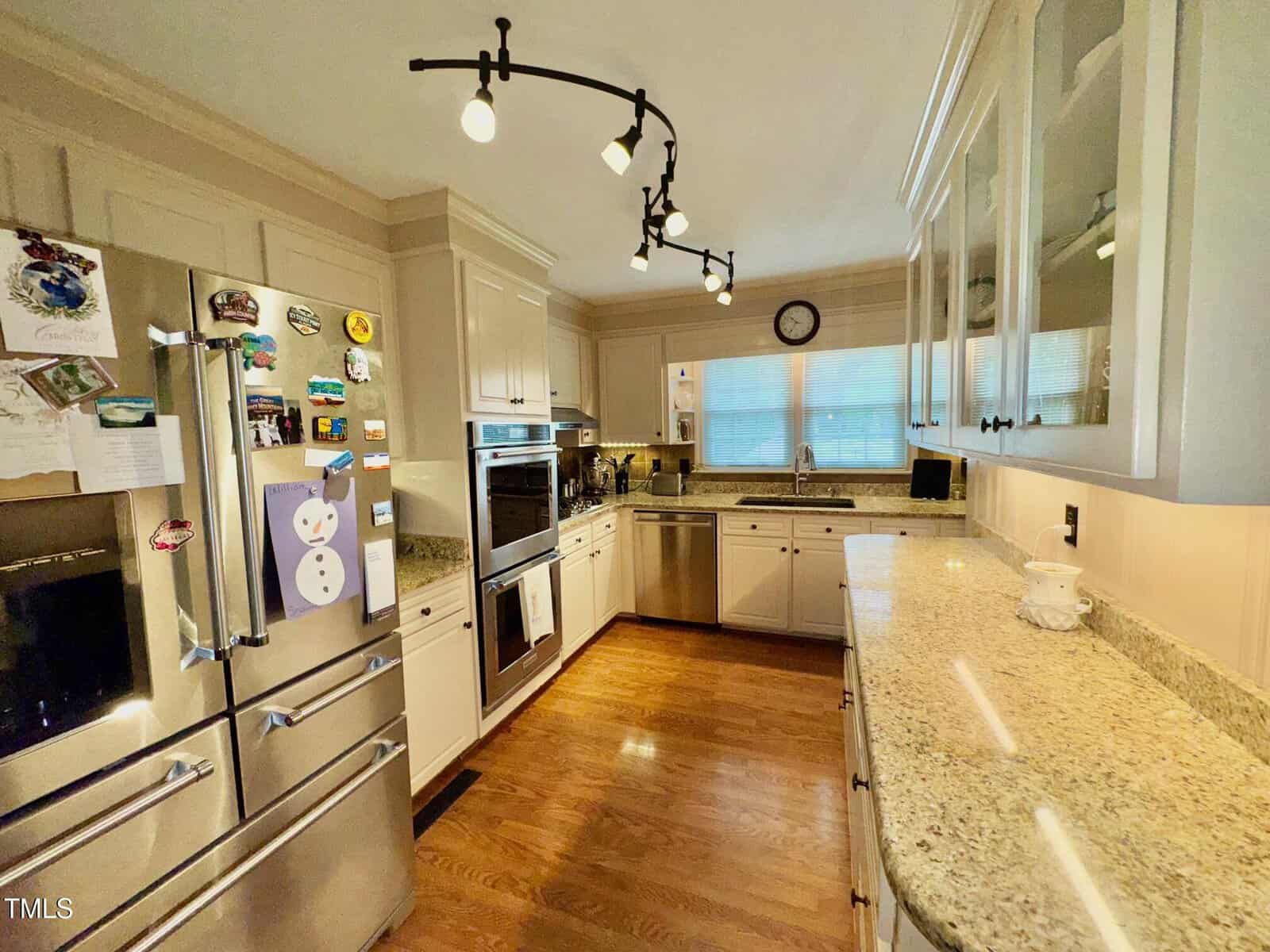 A modern kitchen features stainless steel appliances, including a double oven and refrigerator adorned with magnets. Light wooden cabinets and granite countertops line the walls. A track lighting fixture is mounted on the ceiling. A clock is above the window.