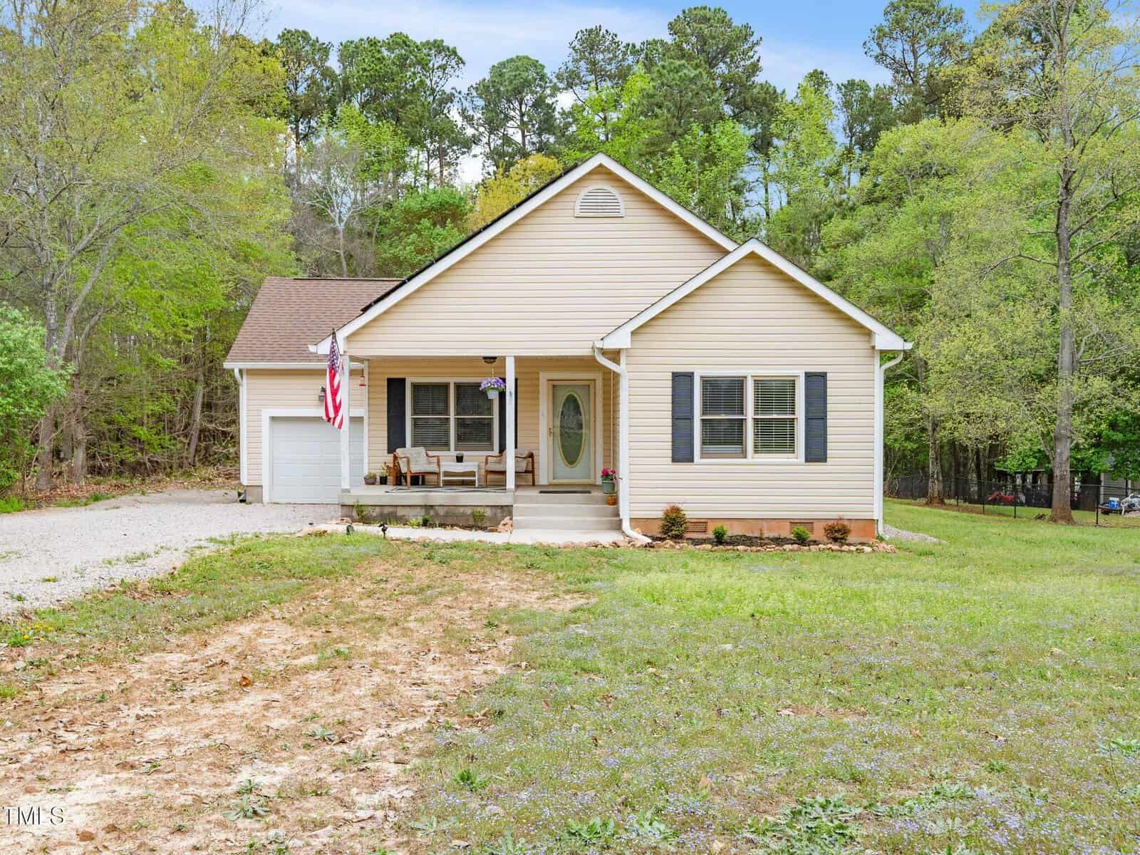 A single-story beige house with dark shutters, a front porch, an American flag, and attached garage, sits on a grassy lot surrounded by tall trees. The driveway is gravel and the lawn has some bare patches.