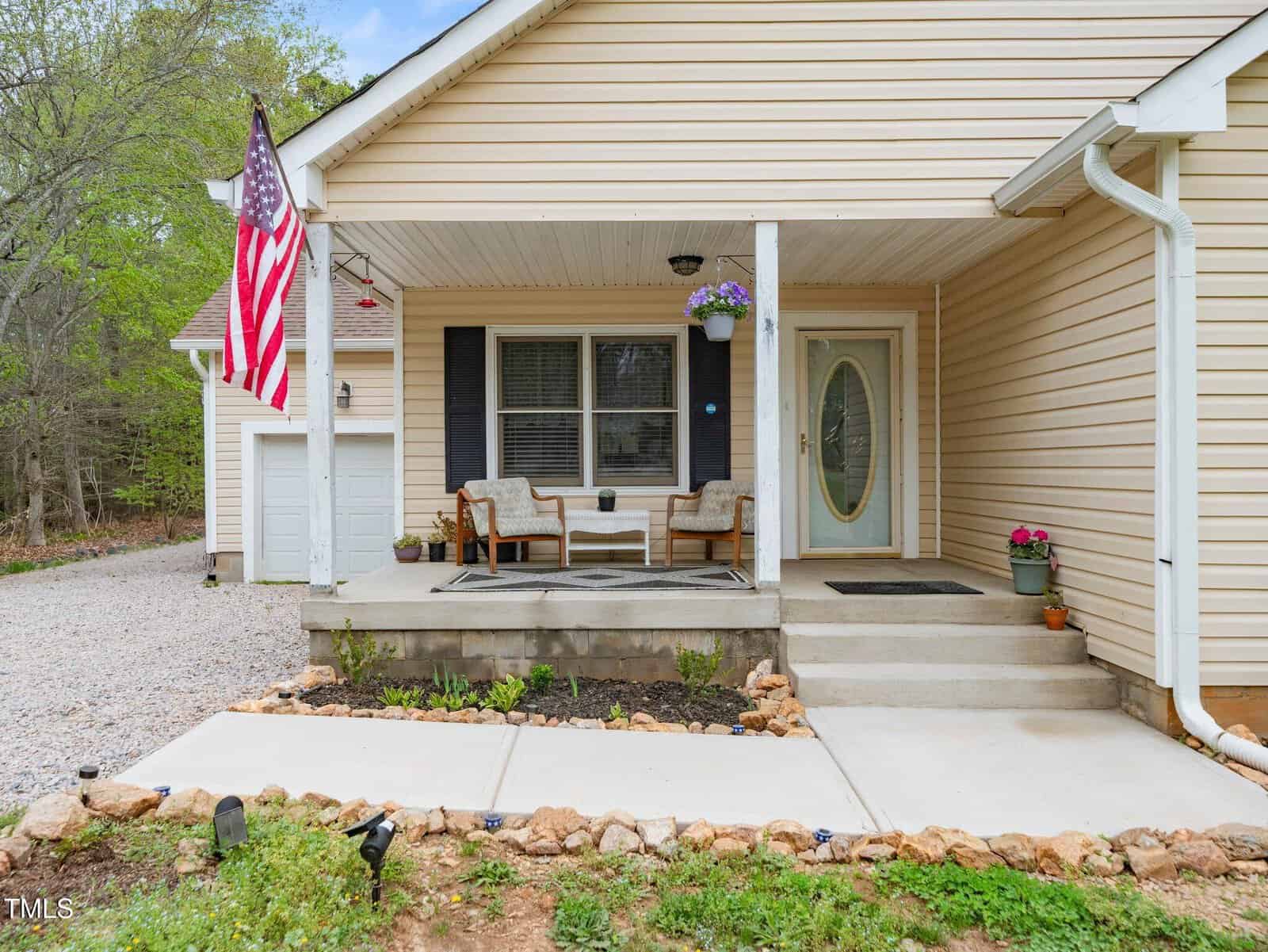 A beige house with a covered front porch featuring two cushioned chairs and a small table. An American flag hangs near the porch. There are potted plants and a gravel driveway on the left side.