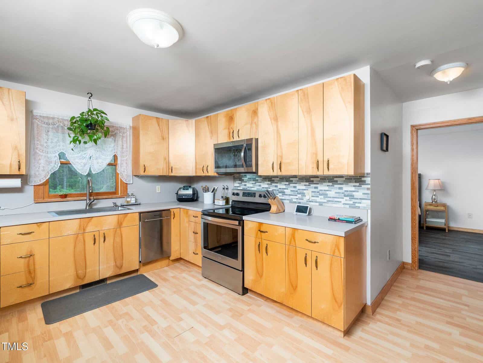 A modern kitchen with light wood cabinets, stainless steel appliances, a tiled backsplash, and light wood flooring. A window with lace curtains and a hanging plant is above the sink. An adjacent room is partially visible.