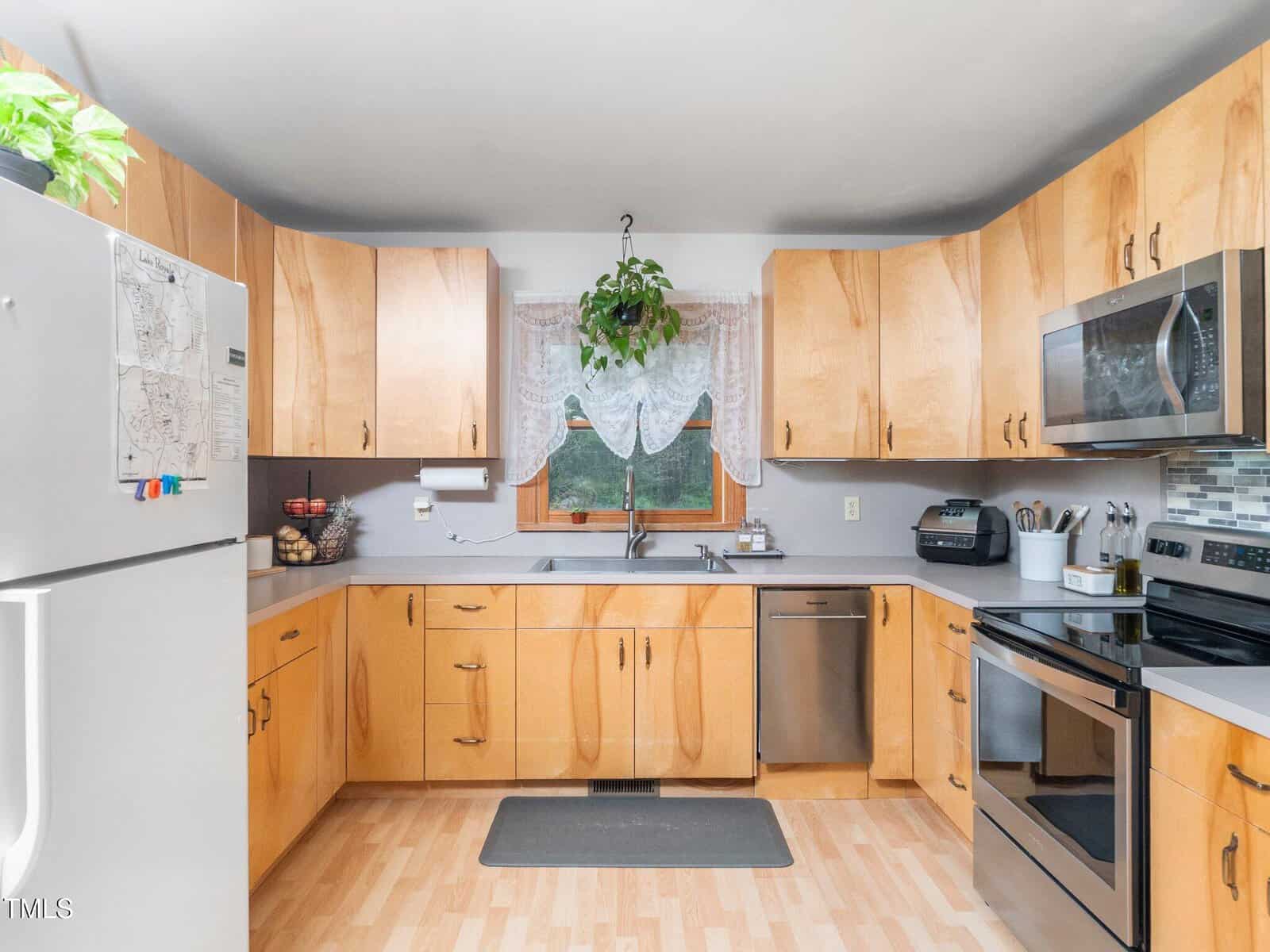 A kitchen with wooden cabinets, stainless steel appliances, a double oven, a dishwasher, a microwave, a window with lace curtains and a hanging plant, and a light wood floor. A refrigerator is on the left.
