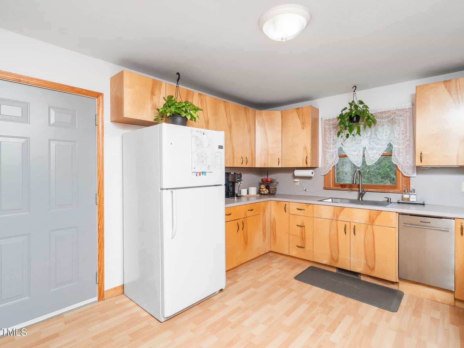 A kitchen with light wood cabinets, a white refrigerator, gray countertop, stainless steel dishwasher, gray door, and two hanging plants near a window with lace curtains. A ceiling light is visible overhead.