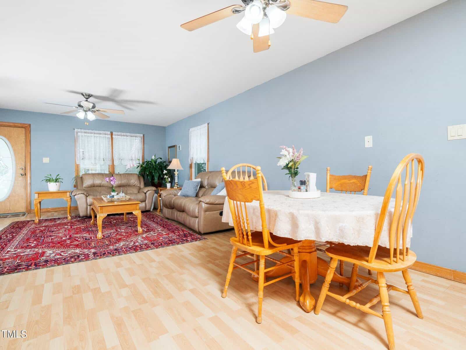 A living and dining area with wood floors, light blue walls, two ceiling fans, a round table with four chairs, two beige sofas, a red rug, plants, and windows with sheer curtains.