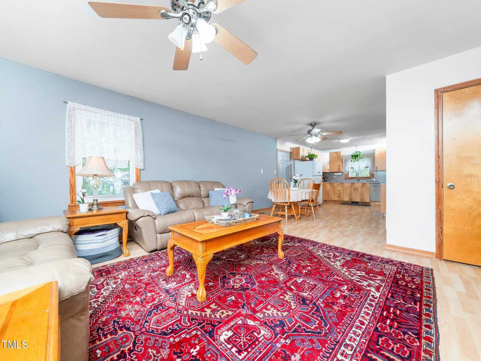 Living room with beige sofas, wooden coffee table, and red patterned rug. Dining area with wood table and chairs is visible in the background, along with the kitchen. Ceiling fans are present in both areas.