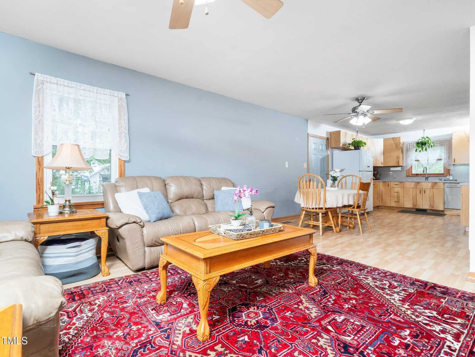 A living room with beige sofas, a wooden coffee table on a red patterned rug, and a side table with a lamp. In the background, there is a dining area with a wooden table and chairs, and a kitchen with light wood cabinets.