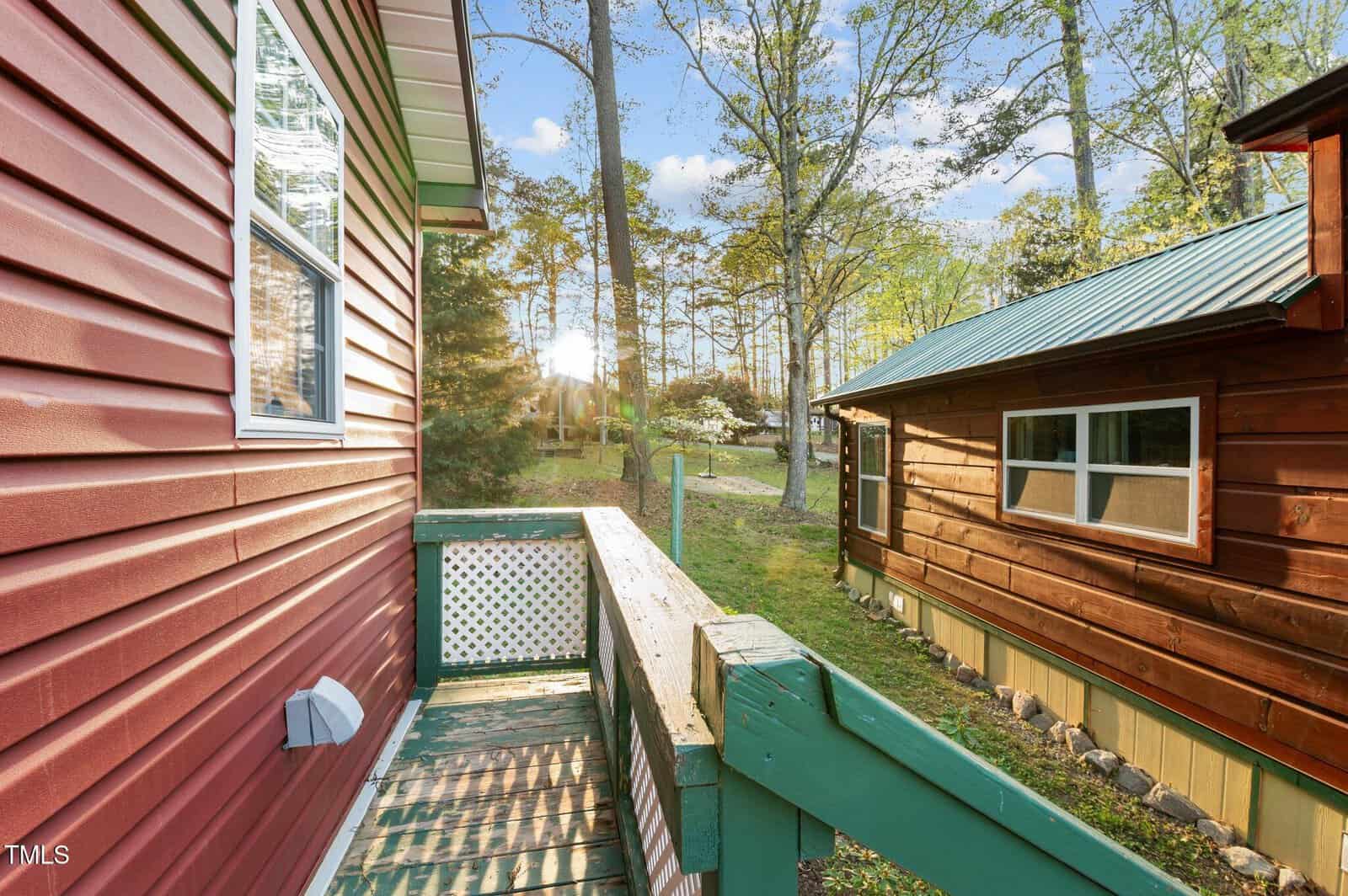 A small deck with green railing connects two wooden cabins. Trees and sunlight are visible in the background, and the ground is covered with grass. The left cabin is red; the right cabin is brown with yellow trim.