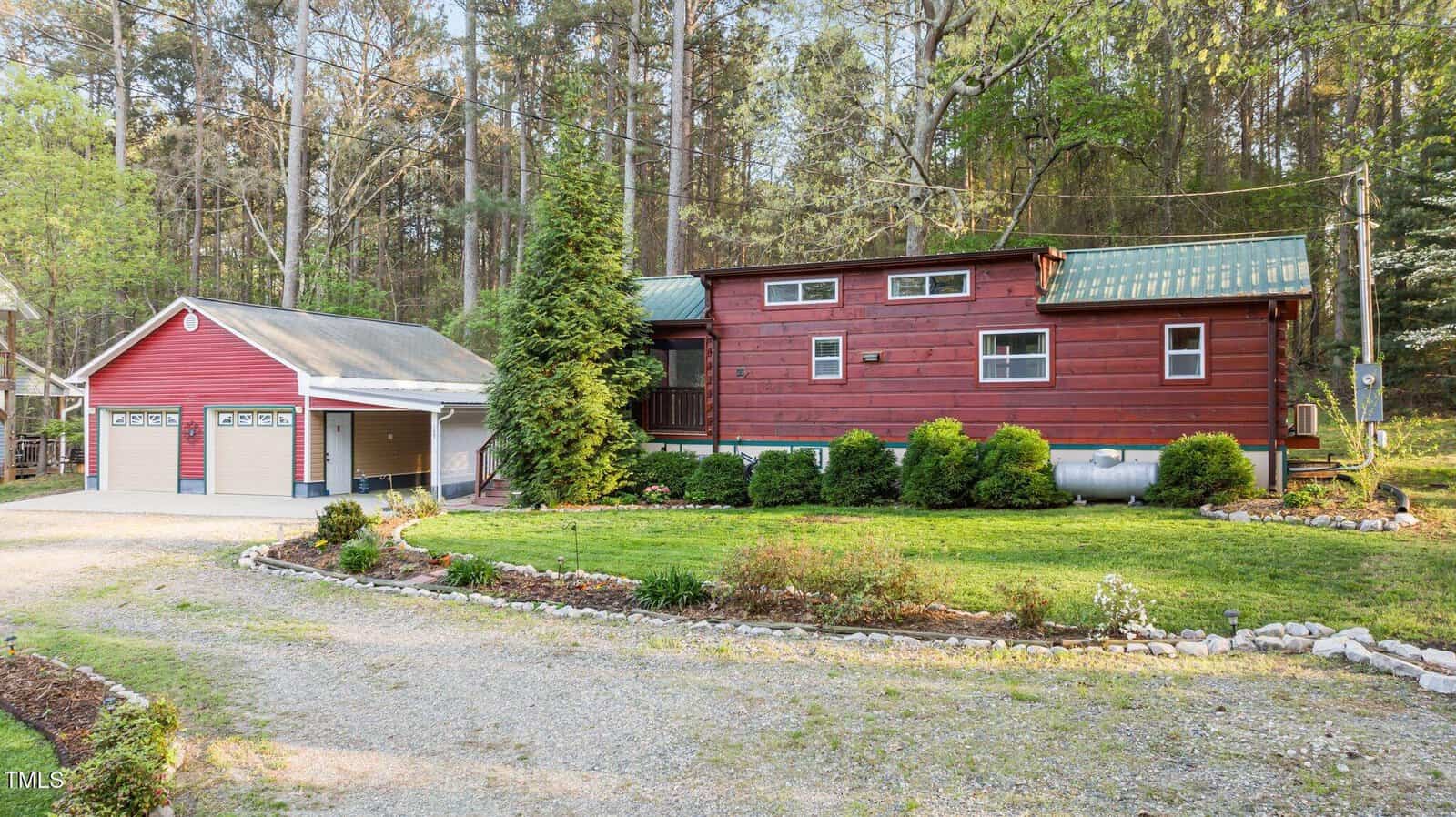 A red log-style house with a green metal roof sits beside a matching detached two-car garage. The property has a gravel driveway, manicured lawn, shrubs, and is surrounded by tall trees and greenery.