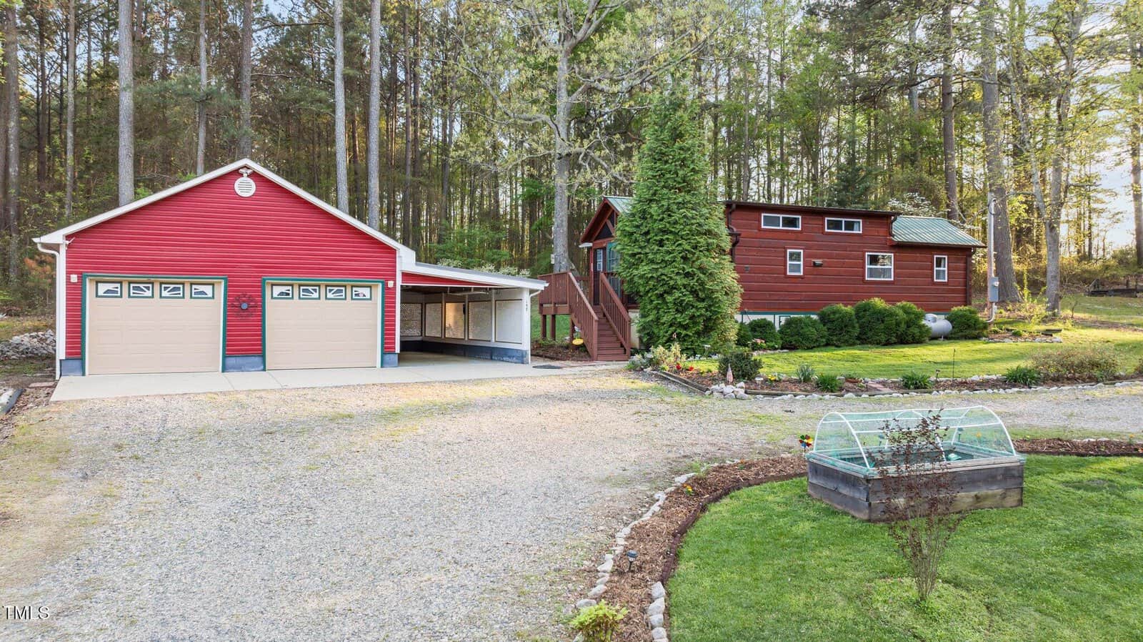 A gravel driveway leads to a red two-car garage with a carport and a small red wooden house with a loft, surrounded by greenery, trees, and a raised garden bed in the foreground.