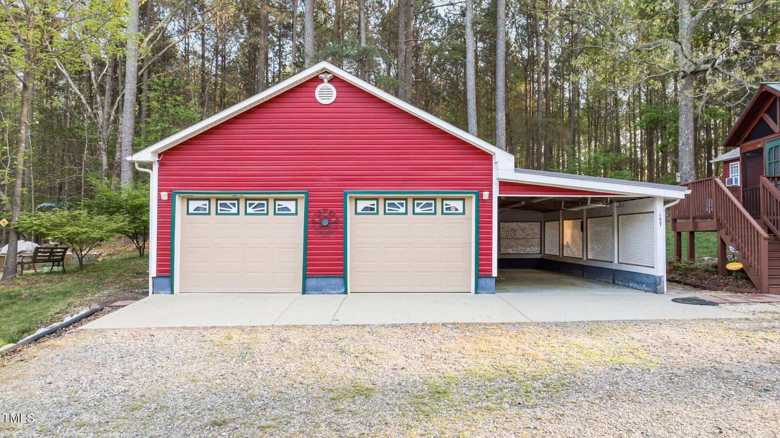 A red building with two beige garage doors and a covered carport stands in front of a wooded area. The structure is surrounded by pavement and gravel, and a staircase is visible to the right.