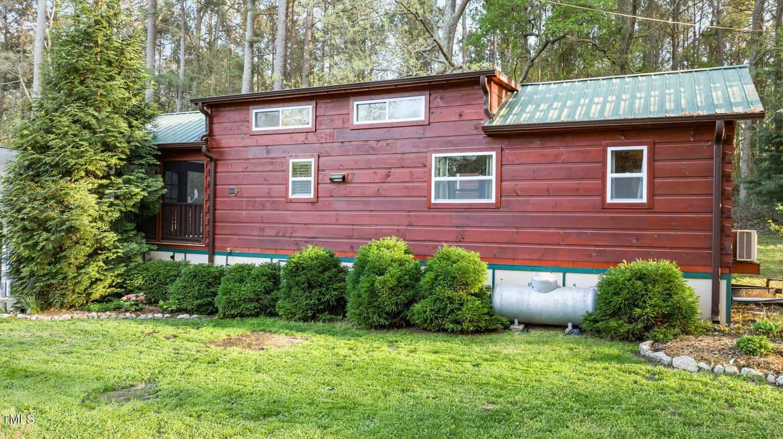 A red-brown wooden tiny house with a green metal roof sits on a grassy yard, surrounded by bushes and a tree, with a propane tank visible near the foundation. Tall trees are in the background.