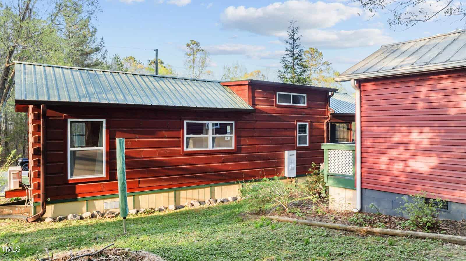 Red wooden tiny house with a metal roof sits on a grassy yard next to another red building. Trees and bushes are in the background under a partly cloudy sky.