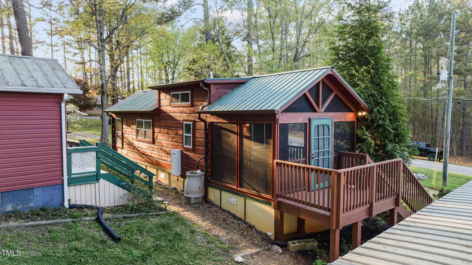 A small wooden cabin with a green metal roof, a screened-in porch, and a raised deck sits next to a purple house surrounded by trees and greenery. A sloped path leads to the road.