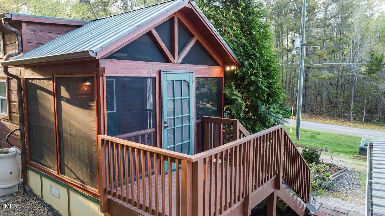 A small screened porch with a green door and wooden railings is attached to a cabin. Stairs lead down to a yard, and a road and trees are visible in the background.