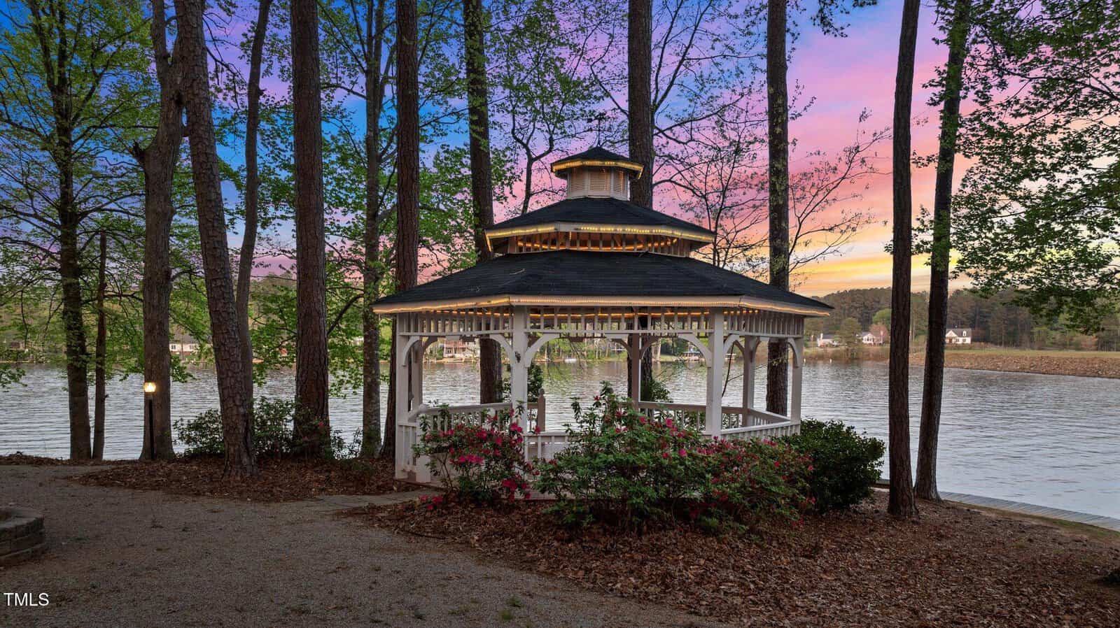 A white gazebo surrounded by trees and shrubs sits near a lake at sunset, with calm water and houses visible across the shore in the background.