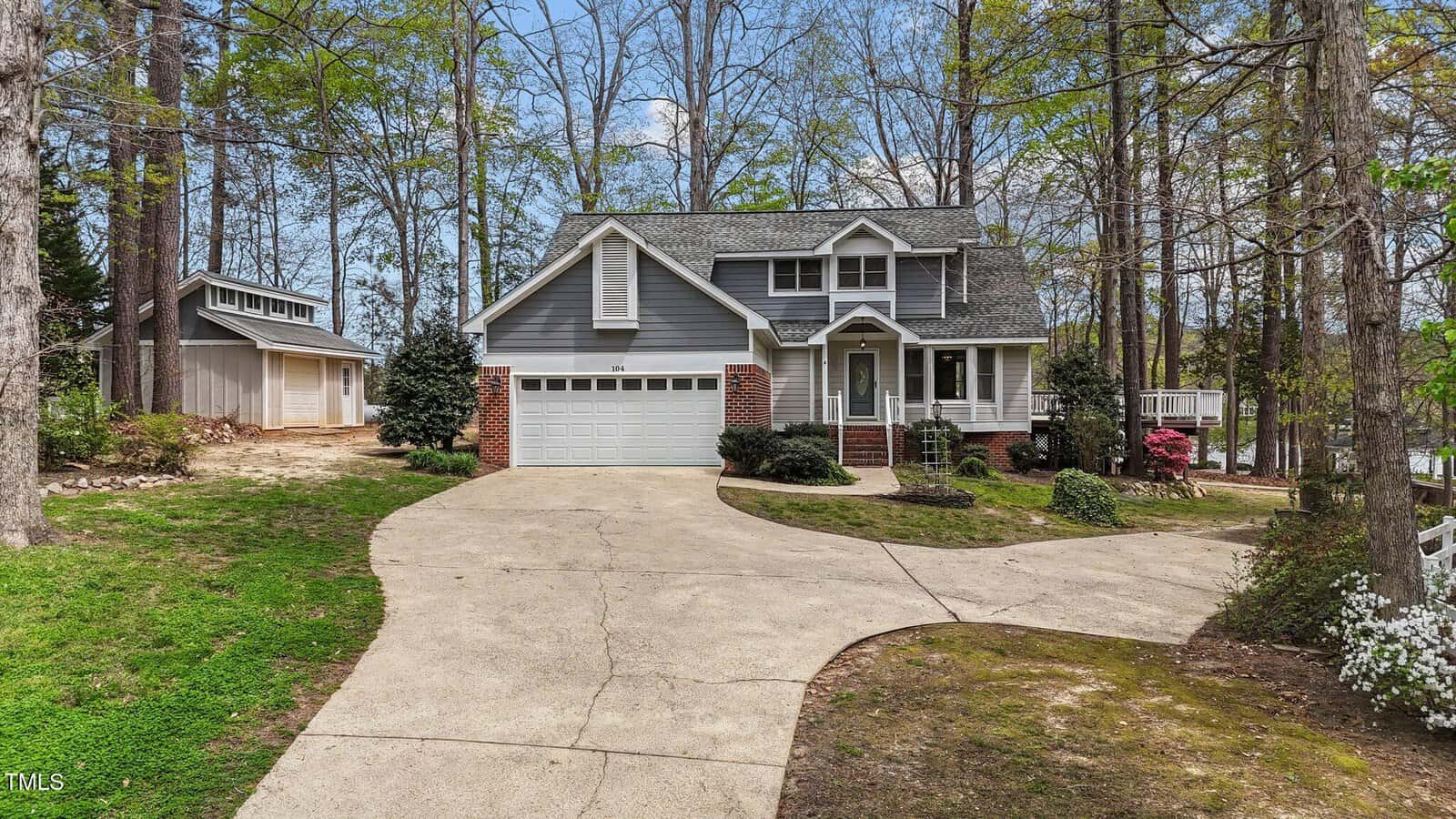 A two-story gray house with white trim, brick accents, and an attached garage sits among tall trees at 104 Stage Line Cove in Louisburg. A detached shed is visible on the left, while curved concrete driveways lead to both structures.