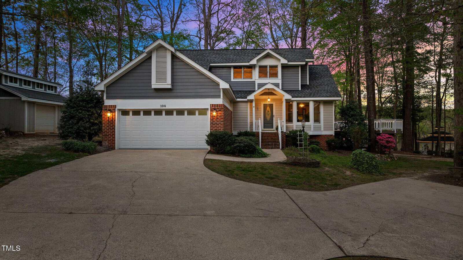 Two-story gray house with white trim, attached two-car garage, brick steps to the front door, and a curved driveway. Surrounded by trees and landscaped bushes, at dusk with a pink and blue sky in the background.