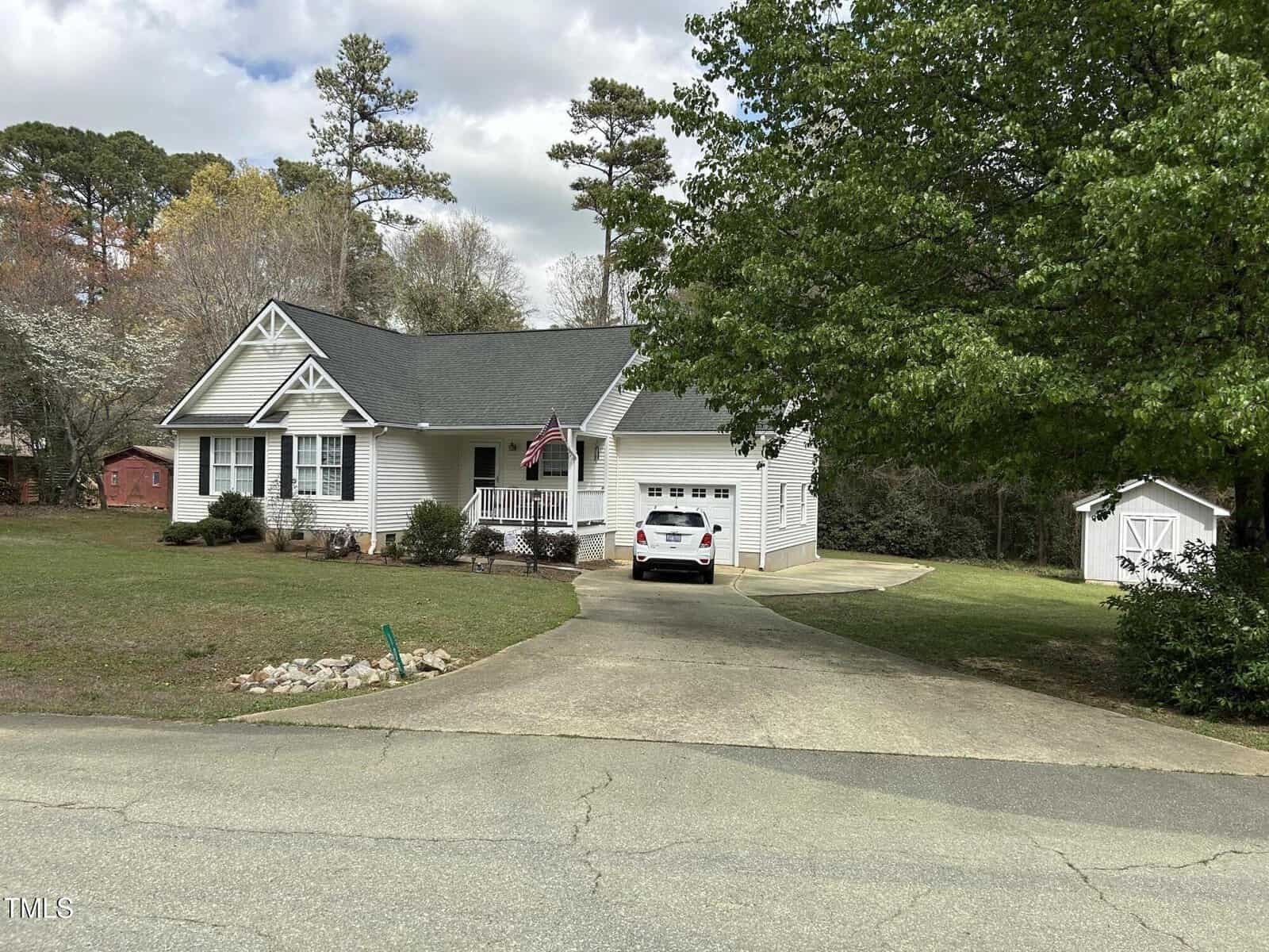 A single-story white house with a front porch, an American flag, and an attached garage is shown. A white car is parked in the driveway, and there is a green lawn with trees and a small white shed in the background.