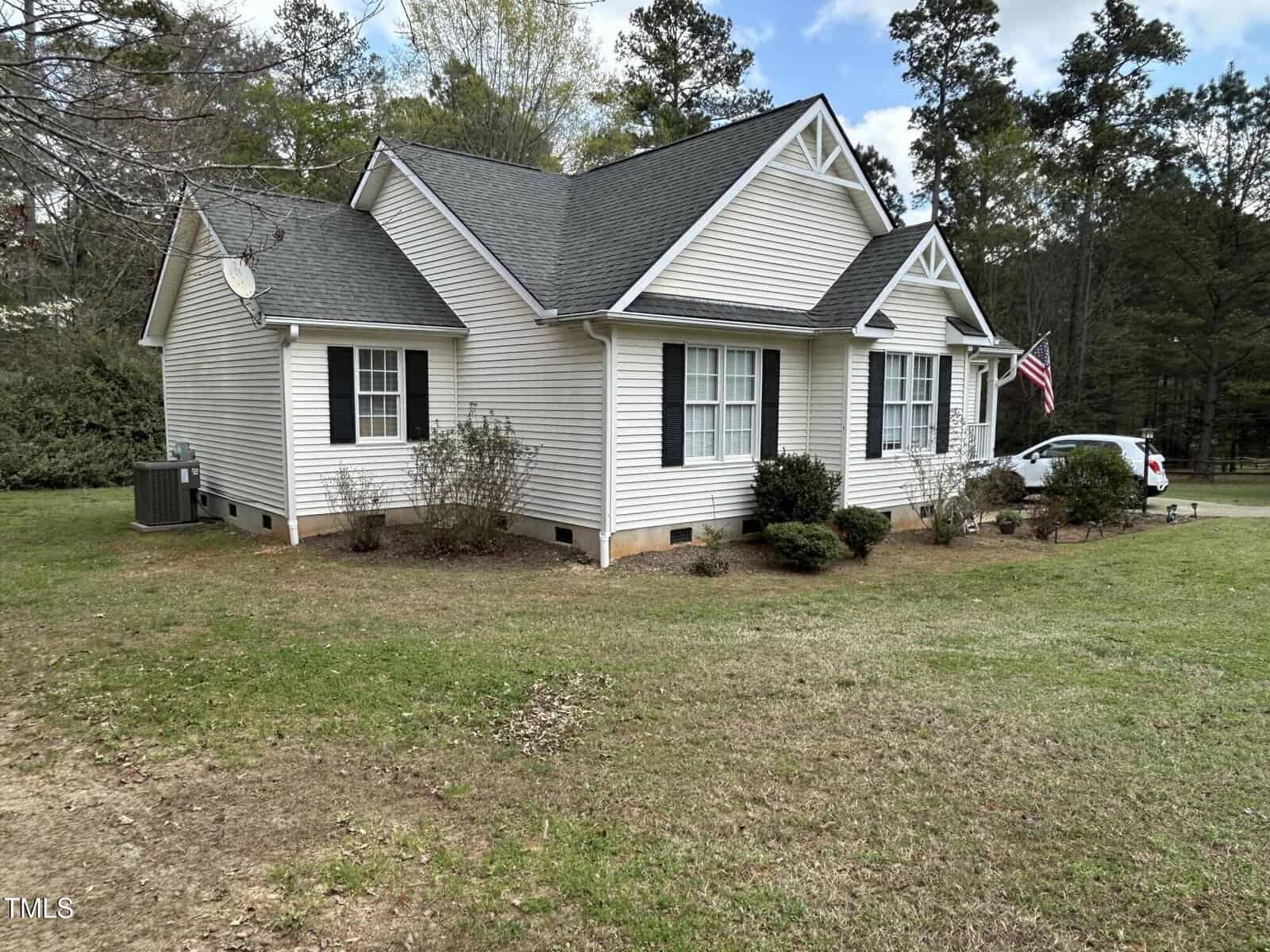 Single-story house with light-colored siding, dark shutters, and an American flag by the front door. The yard has patchy grass and some bushes near the foundation. A white car is parked in the driveway.