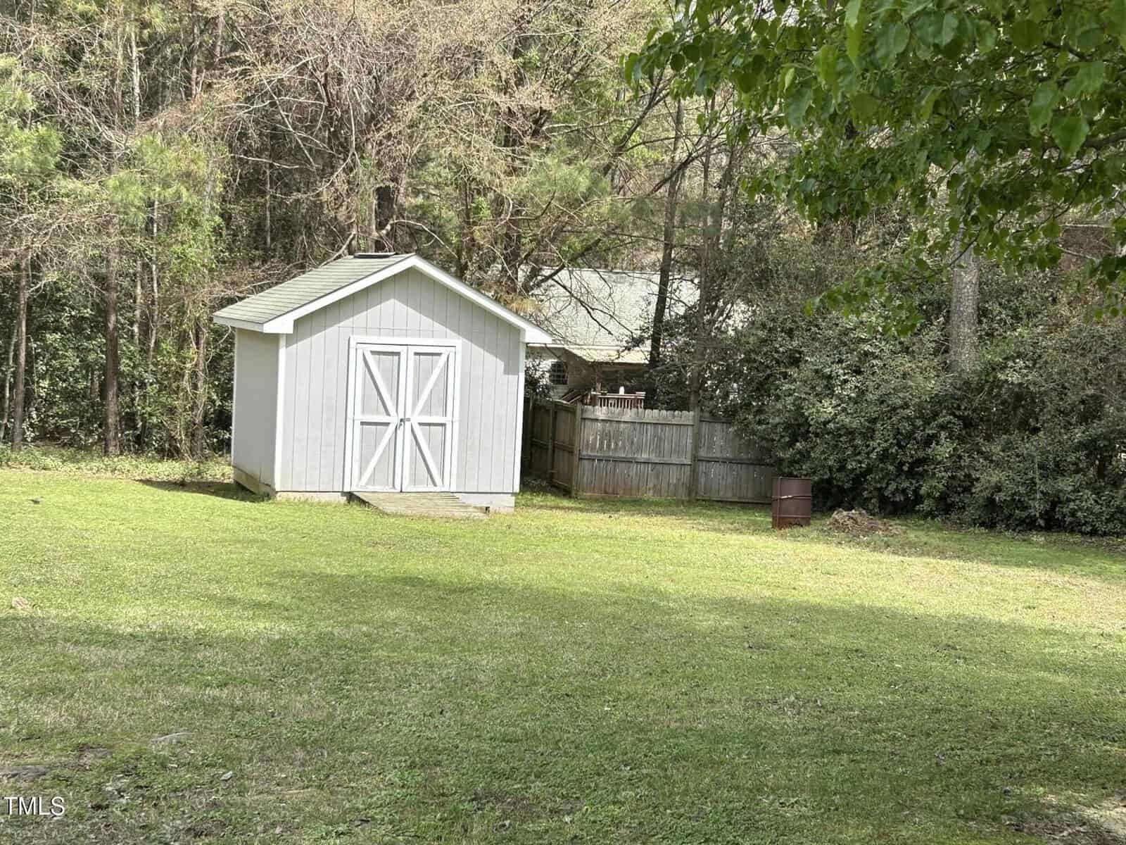 A small white shed with double doors sits on a grassy lawn near a wooden fence, surrounded by trees and shrubs on a sunny day.